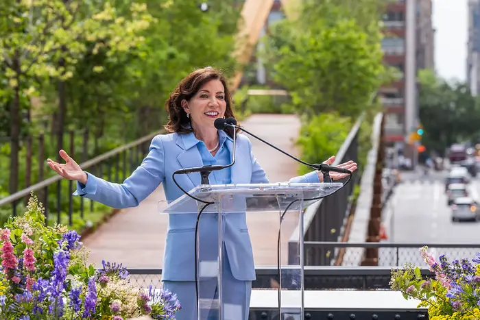 Gov. Kathy Hochul, in a powder blue suit, stands behind a clear podium on the High Line surrounded by trees and flowers.